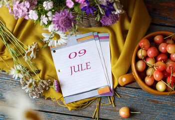 Calendar for July 9: flip calendar with numbers 09, name of the month July in English on a yellow napkin with wild flowers and ripe cherries