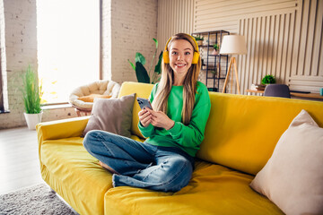 Smiling young woman in a green sweatshirt relaxing on a yellow sofa holding a smartphone in a cozy...