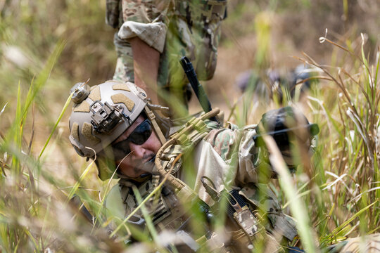 Special forces soldier holding rifle and communicating on radio during tactical operation in forested mountain terrain with full combat gear and camouflage uniform.