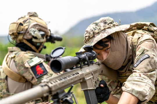 Sniper aiming through rifle scope in military training, close-up on open lens cap, with blurred soldier in background wearing camouflage and tactical gear. - Powered by Adobe