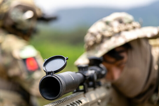 Sniper aiming through rifle scope in military training, close-up on open lens cap, with blurred soldier in background wearing camouflage and tactical gear.