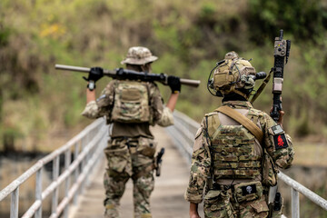 Two armed special forces soldiers in full tactical gear patrolling a narrow bridge during field...