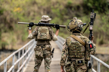Two armed special forces soldiers in full tactical gear patrolling a narrow bridge during field...
