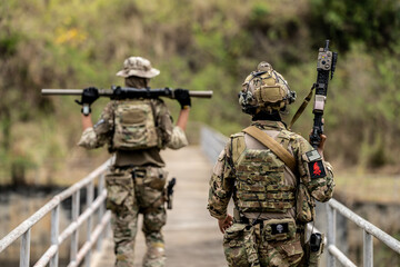 Two armed special forces soldiers in full tactical gear patrolling a narrow bridge during field...