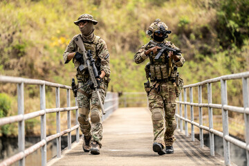 Two armed special forces soldiers in full tactical gear patrolling a narrow bridge during field...