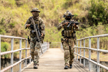 Two armed special forces soldiers in full tactical gear patrolling a narrow bridge during field...
