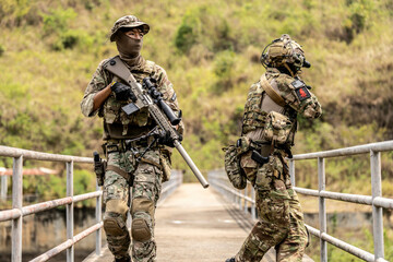 Two armed special forces soldiers in full tactical gear patrolling a narrow bridge during field...