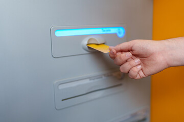Hand inserting a gold credit card into an illuminated ATM cash machine, glowing with blue light,...