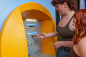 Young women using credit card, withdrawing cash from ATM while standing together on sunny street corner