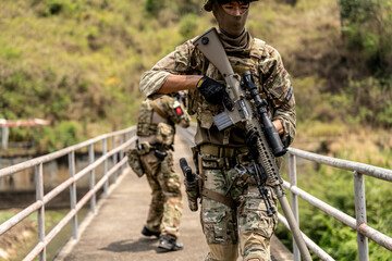 Two armed special forces soldiers in full tactical gear patrolling a narrow bridge during field...