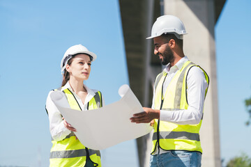 Group of construction professionals at job site outdoors inspecting blueprints, discussing technical layouts and coordinating development