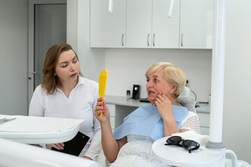 Senior female patient with dentist looks in the mirror and having dental treatment at dentist's...