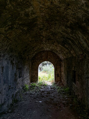 A dark and weathered stone tunnel with a vaulted ceiling opens into a sunlit clearing. The arched...