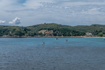 A tranquil scene of sailboats anchored in a calm bay, surrounded by lush green hills on a partly cloudy day. The turquoise and deep blue hues of the water create a striking contrast with the forest.