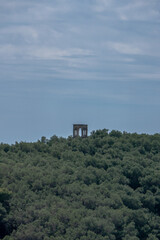 A solitary ancient stone pavilion with arched openings standing atop a lush green forested hill, under a partly cloudy blue sky.
