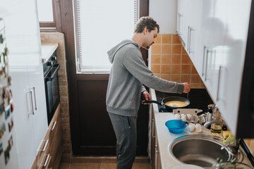 A man is seen cooking in a modern home kitchen, preparing eggs on a stove top while wearing casual...