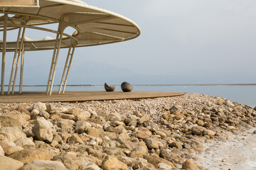 Obraz na płótnie Canvas Minimalist view of Dead Sea beach with modern canopy structure, stony shore and tranquil saltwater. Tranquil landscape for travel, spa or wellness concept with copy space and natural tones.