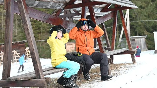 A heartwarming scene of a man and child, bundled in warm winter gear, resting on a wooden bench after a day of skiing. The snowy winter landscape surrounds them.