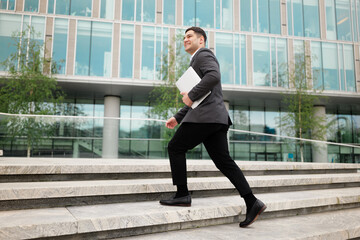 Business professional walking confidently up steps with laptop in hand toward a modern office...