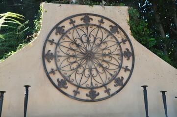 Decorative Iron Circle on Stucco Wall Surrounded by Foliage – Scrollwork with Classic Fleur-de-Lis Details in Sunlight
