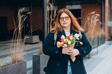 Beautiful young woman with spring tulips flowers bouquet walking at city street. Happy girl posing outdoors. Spring portrait of pretty female on urban background. High quality photo.