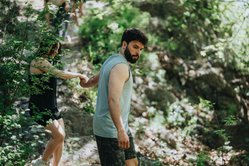 One person assists another as they walk through a shaded forest trail, highlighting exploration,...