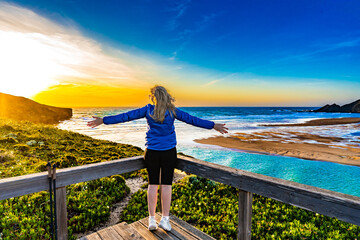 Beautiful middle aged woman standing with outstretched arms at viewpoint on picturesque Amoreira beach on Algarve coast in Portugal on sunrise. Back view
