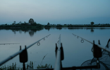 Obraz premium Fishing rods with bite alarms set up by a tranquil body of water, hinting at a peaceful day of carp fishing.