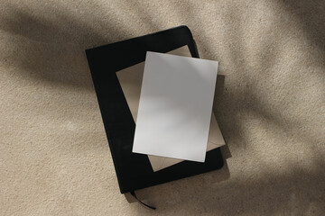 Blank white greeting card, invitation mockup on black notebook. Kraft envelope placed on sandstone...