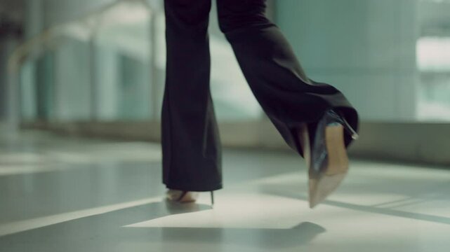 A woman wearing black pants and high heels is walking down a hallway. She is wearing a black dress and has her legs spread apart