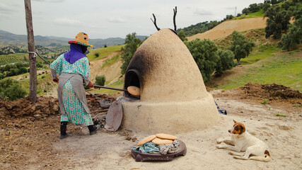 Taounate, Morocco - Mai, 04, 2025 : A Berber woman removes freshly baked traditional bread from an earthen oven in the green mountains of Taounate, Morocco.