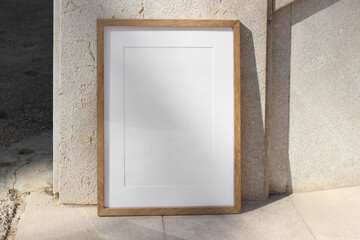 Fototapeta na wymiar Vertical wooden picture frame leaning against stone wall in sunlight. Textured beige tiles background. Summer outdoor natural, minimal backdrop. Empty poster mockup, branding display.
