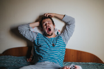 A young man yawning while sitting in bed, wearing pajamas. The image captures the feeling of...