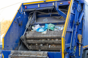 Garbage truck rear loading trash compactor filled with various waste items including plastic and paper during daytime street collection