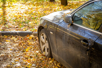 Damaged black car in autumn setting, front left view, dent on side, scattered fallen leaves, parked on leaf-covered ground, afternoon sunlight, no people, nature background