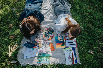 Two teenage girls lying on a blanket outdoors creating paintings with vibrant colors, surrounded by...