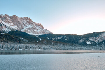 Breathtaking Alpine Winter Wonderland: Majestic Zugspitze Mountain Reflects in Frozen Eibsee Lake Amidst Snowy German Landscape Picturesque Ski Resort Nestled in Bavarian Forest Offers Stunning