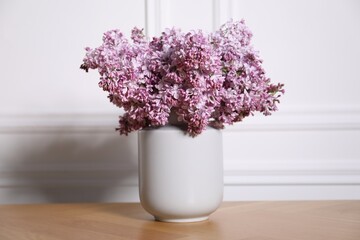 Beautiful lilac flowers in vase on wooden table near white wall indoors