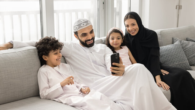 Joyful family leisure time. Happy smiling Muslim couple with diverse junior school age kids daughter son wear national Arabic clothes watch funny video online via smartphone on couch at living room