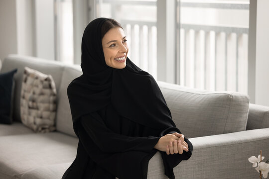 Modern Muslim elegance. Portrait happy beautiful young Arabian woman wear traditional Islamic dress abaya hijab relax on comfy couch look aside with shiny smile enjoy peaceful moment of home comfort - Powered by Adobe