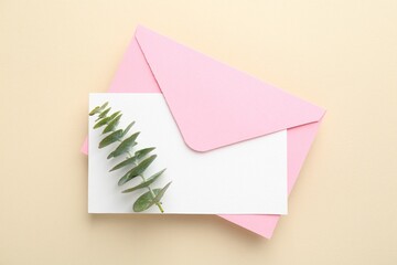 Blank invitation card, envelope and eucalyptus branch on beige background, top view