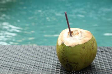 Green coconut with refreshing drink and straw on wicker table near swimming pool outdoors, closeup....