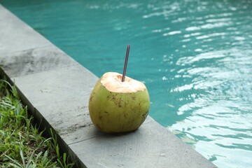 Green coconut with refreshing drink and straw near swimming pool outdoors