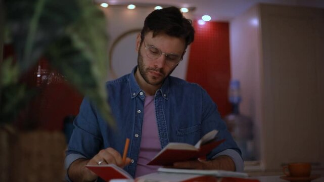 Focused young man in glasses writing in a notebook while reading, seated in a cozy kitchen. Evening scene with soft lighting, capturing study, planning, or journaling at home.
