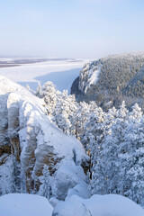 Snow-covered cliffs and evergreen trees overlook a frozen river, creating a serene winter landscape...