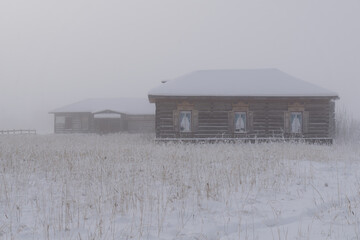 Snow-covered wooden houses in a foggy landscape, surrounded by tall grass, creating a serene winter...