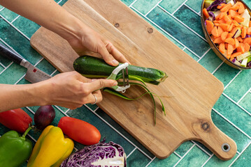 Close-up of a female hand using a vegetable peeler to remove the skin of a fresh zucchini. Surrounded by raw vegetables and green vintage-style tiles, the scene evokes healthy eating, mindful cooking,