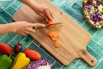 Woman’s hand slicing a vibrant orange carrot with a sharp knife in a home kitchen. Surrounded by other colorful vegetables and a green tile wall, this cozy scene reflects healthy eating and homemade.