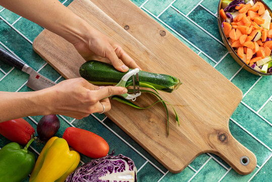 Close-up of a female hand using a vegetable peeler to remove the skin of a fresh zucchini. Surrounded by raw vegetables and green vintage-style tiles, the scene evokes healthy eating, mindful cooking,