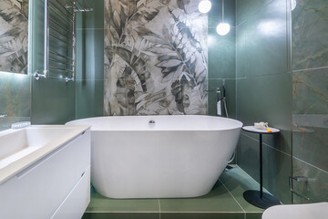 A contemporary bathroom showcases a spacious freestanding bathtub against a tropical-themed wall. Stylish lighting and modern design elements enhance the relaxing atmosphere.